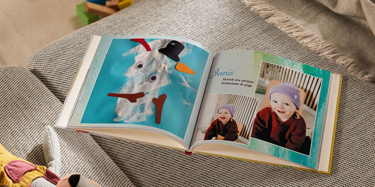 Sur un sofa, un LIVRE PHOTO CEWE ouvert est posé à côté d’un animal en peluche et d’une couverture. La page de gauche montre la photo d’un bricolage d’enfant représentant un bonhomme de neige. Sur la page de droite, on voit deux photos d’un enfant qui rit. Le titre mentionne « Janis bricole son premier bonhomme de neige ». L’arrière-plan de la double page est rempli d’aquarelles bleues et vertes.
