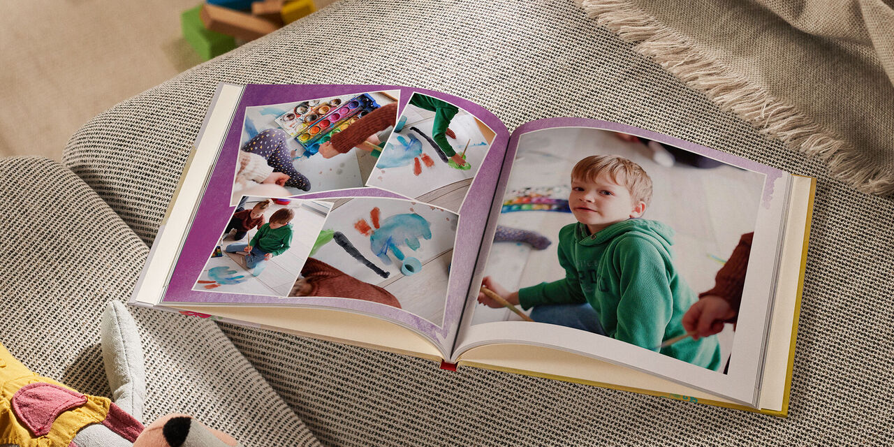 Un LIVRE PHOTO CEWE ouvert est posé sur un canapé à côté d’un animal en peluche et d’une couverture. Sur la page de gauche, on distingue quatre photos qui se chevauchent légèrement. Toutes montrent un enfant en train de faire un dessin avec les couleurs d’une boîte à encre. La page de droite montre une grande photo d’un garçon qui regarde l’appareil photo. La double page est surlignée d’un motif d’aquarelle violet.