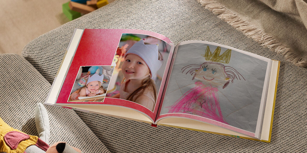 Un FOTOLIBRO CEWE aperto si trova su un divano accanto a un peluche e a una coperta. La pagina di sinistra mostra due foto di una bambina sorridente che indossa una corona. La pagina di destra mostra il disegno di una principessa che indossa una corona. Lo sfondo della doppia pagina è disegnato con un motivo ad acquerello rosso.